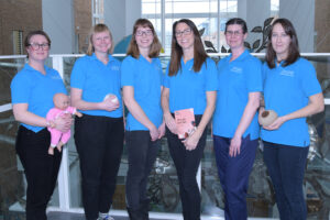 A team photo of six breastfeeding peer supporters. One holds a baby doll. 