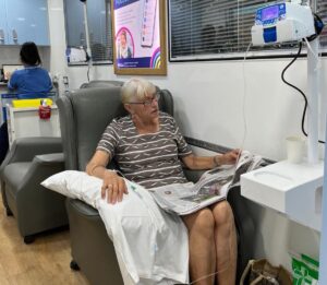 Patient Anne Steel sitting in a chemotherapy chair having treatment. She is reading a paper.